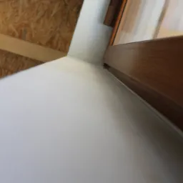 Gładka, biała ściana po nałożeniu tynku maszynowego, widoczny fragment drewnianej ramy okiennej i sufit z płyt OSB.