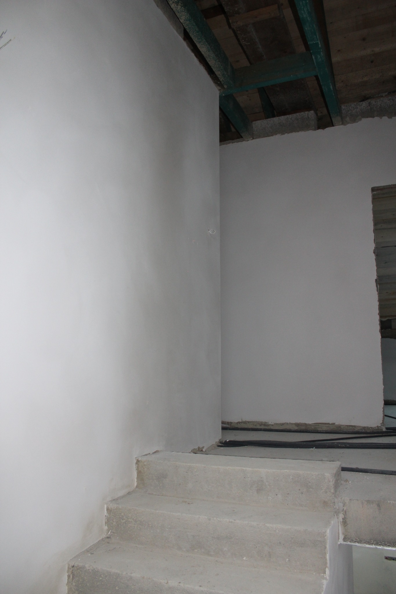 Świeżo otynkowane ściany wewnętrzne z widocznymi surowymi schodami betonowymi i fragmentem drewnianego stropu, ukazujące etap wykończenia wnętrza.