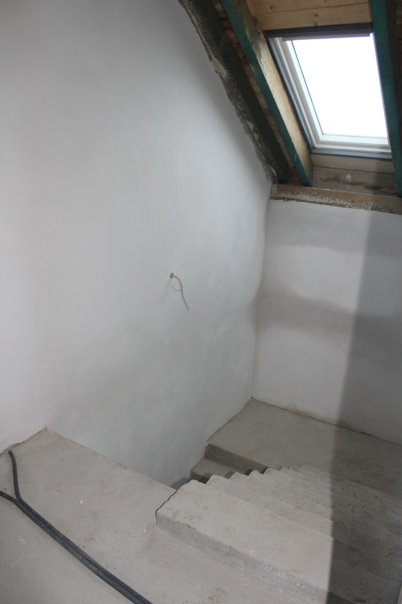Świeżo otynkowane poddasze ze schodami i oknem dachowym; widoczny kabel elektryczny przy podłodze, jasne, gładkie ściany.