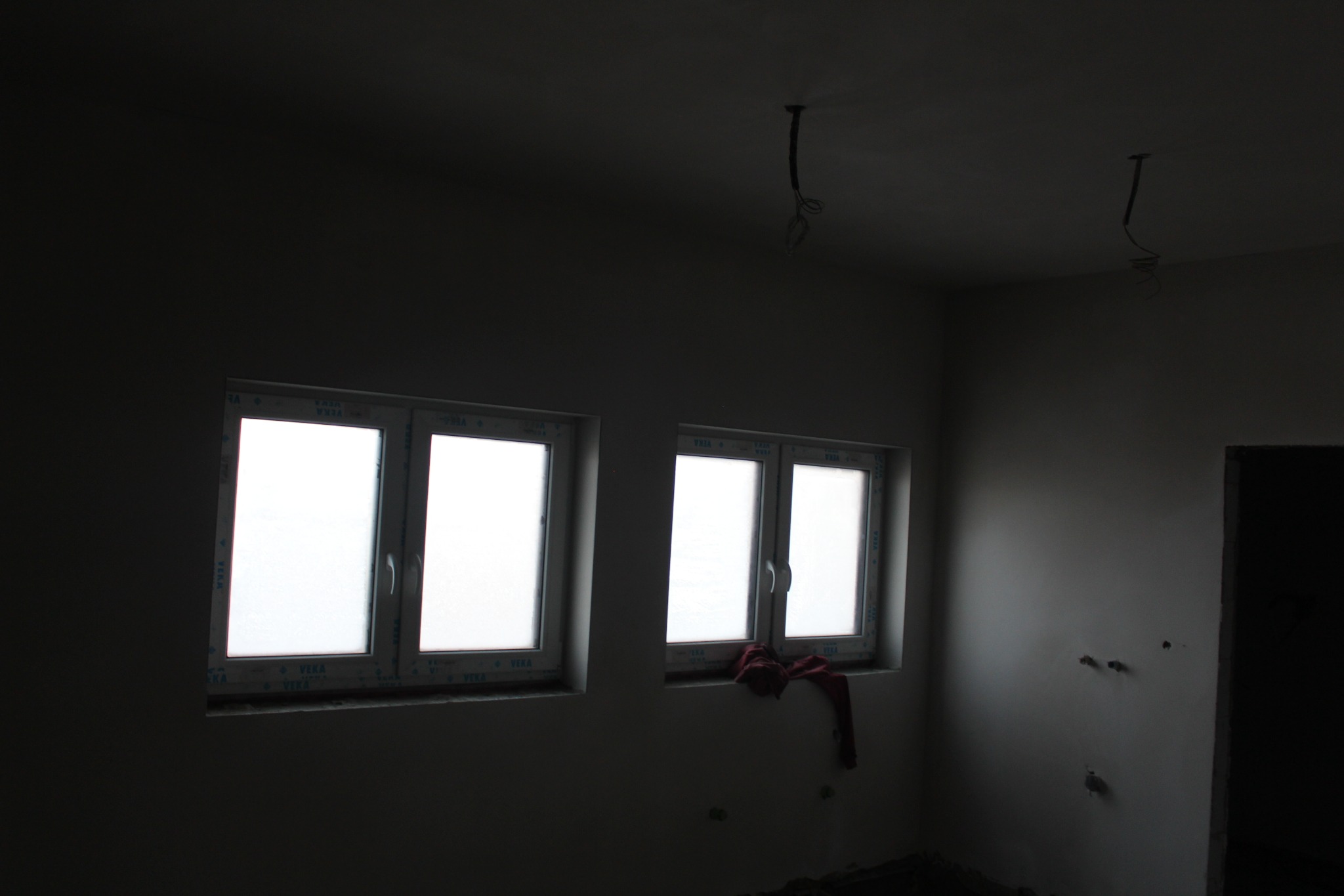 Wnętrze pokoju w trakcie remontu: dwie białe okna PCV, świeżo otynkowane ściany w odcieniach szarości, wystające kable z sufitu, czerwona szmatka na parapecie.