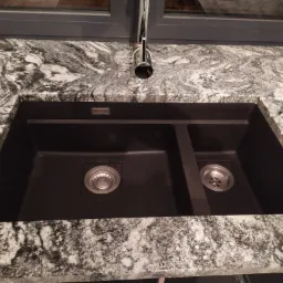 Czarny, dwukomorowy zlew wpuszczony w blat z szarego, wzorzystego kamienia, bateria chromowana, widok z góry.