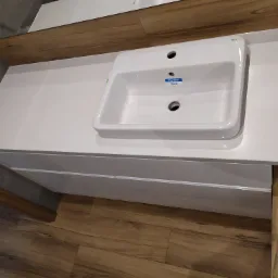 Nowoczesna, biała umywalka Roca The Gap na białym blacie z szafką w łazience z drewnopodobnymi elementami wykończeniowymi.