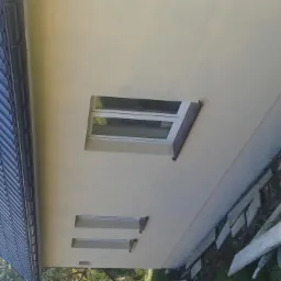 Ściana domu po ociepleniu z dwoma małymi oknami i jednym dużym oknem dachowym, widoczne deski leżące przy fundamencie.