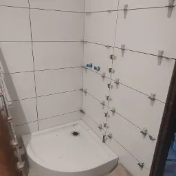 Białe, kwadratowe płytki z krzyżykami dystansowymi na ścianie w rogu łazienki. Widoczny brodzik i fragment drabiny. Etap prac wykończeniowych.