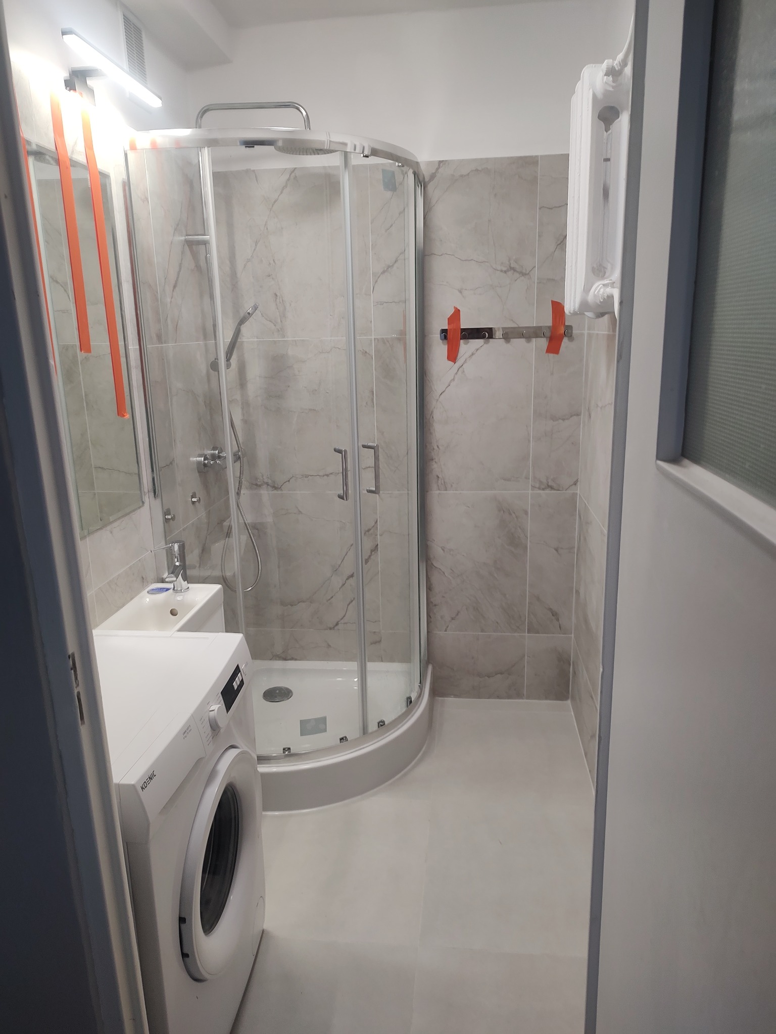 Jasna łazienka z kabiną prysznicową, umywalką i pralką w jednym. Ściany wyłożone płytkami imitującymi marmur. Widoczny grzejnik i akcesoria łazienkowe.