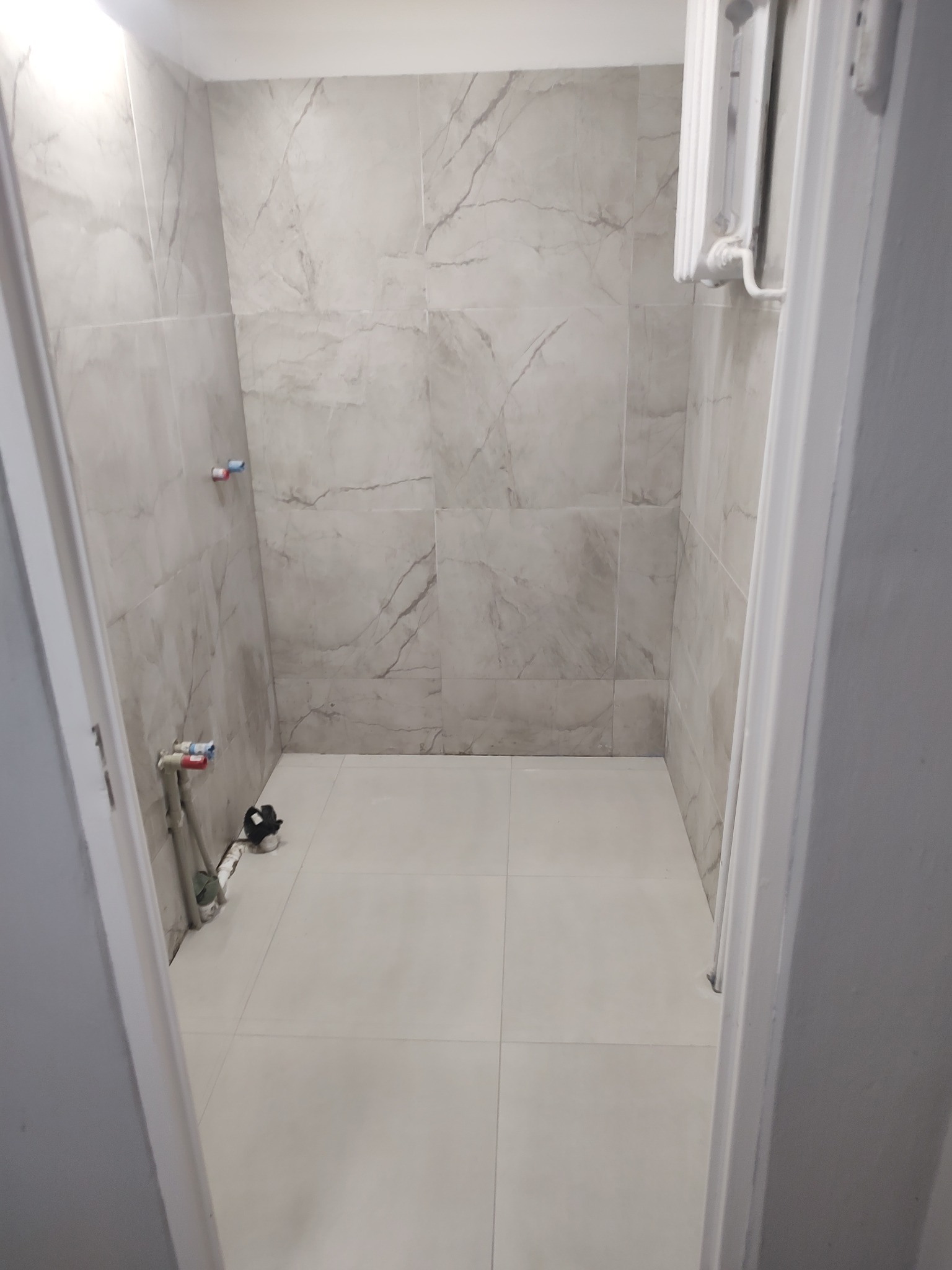 Wykończona łazienka z szarymi płytkami imitującymi marmur na ścianach i podłodze. Widoczne rury i grzejnik. Ujęcie z perspektywy wejścia.