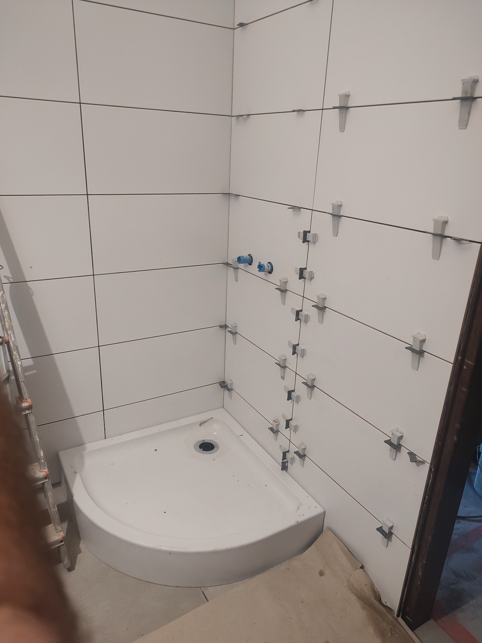 Białe, kwadratowe płytki z krzyżykami dystansowymi na ścianie w rogu łazienki. Widoczny brodzik i fragment drabiny. Etap prac wykończeniowych.