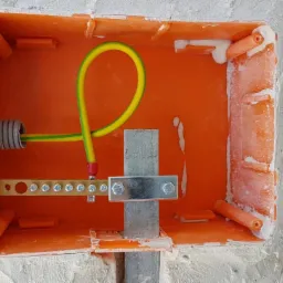 Pomarańczowa puszka instalacyjna w surowej ścianie z widoczną szyną wyrównawczą potencjałów i przewodem ochronnym żółto-zielonym.
