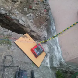 Miernik elektryczny leżący na desce obok wykopu pod fundamentem budynku, widoczne przewody i kabel uziemiający oraz wiertarka udarowa.