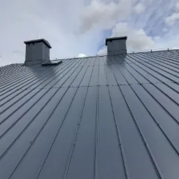 Nowy, szary, blaszany dach z dwoma kominami na tle pochmurnego nieba.