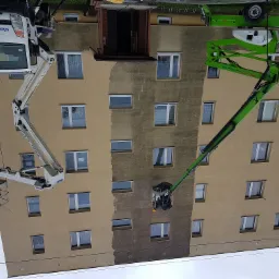 Wielopiętrowy budynek w trakcie odświeżania elewacji, widoczne dwie platformy podnośnikowe z pracownikami, jedna biała i jedna zielona, fragment elewacji po lewej stronie w nowym kolorze, prawa...