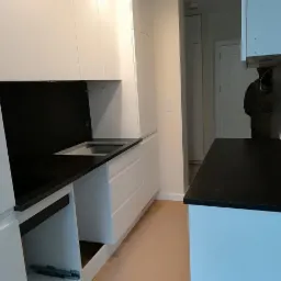 Nowoczesna kuchnia z białymi szafkami i czarnym blatem, widoczne niedokończone szuflady i osoba w tle.