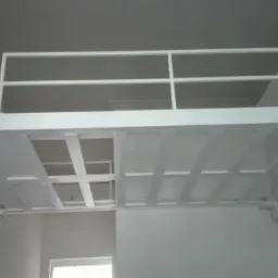 Biała, metalowa konstrukcja antresoli z barierką, zamontowana w pomieszczeniu o białych ścianach i suficie. Widok od dołu, ukazujący sposób mocowania do ścian.