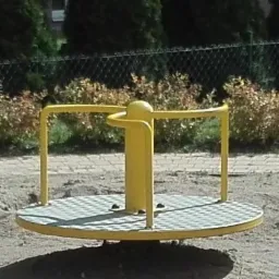 Żółta karuzela tarczowa na placu zabaw, z metalowymi uchwytami i kratownicową powierzchnią do stania, otoczona piaskiem i zielenią.