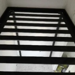 Czarna metalowa konstrukcja z poziomymi szczeblami, prawdopodobnie fragment bramy lub ogrodzenia, widziana od dołu pod kątem, z widocznym fragmentem pomieszczenia w tle.