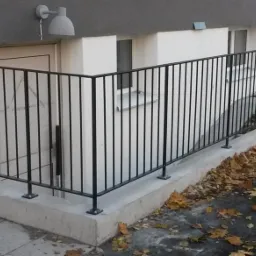 Czarne, metalowe barierki zabezpieczające zejście do piwnicy budynku, widoczne mocowania do betonowej podstawy, jesienne liście na chodniku.