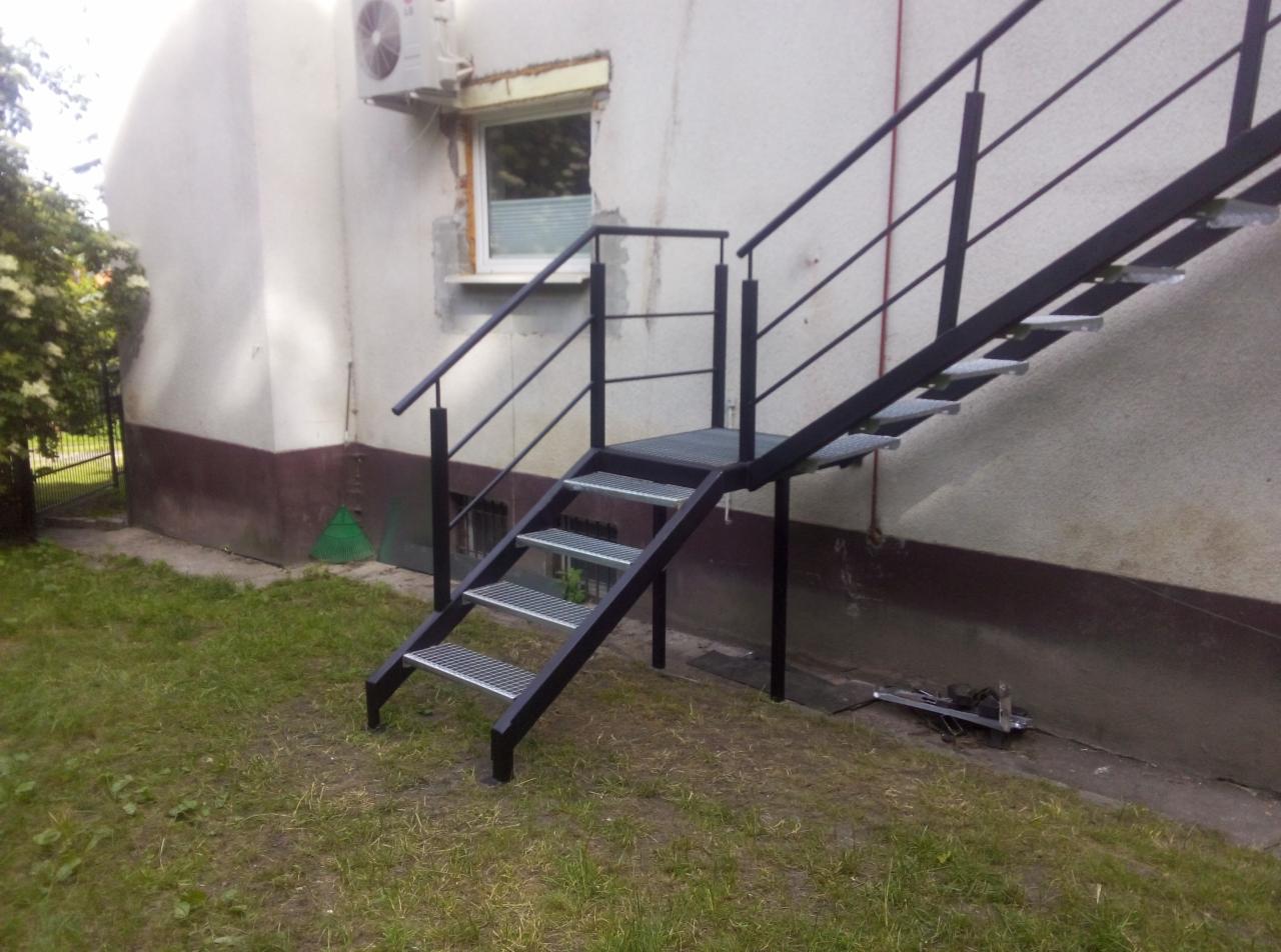 Zewnętrzne metalowe schody z kratowymi stopniami, prowadzące do budynku z klimatyzacją i oknem, widoczne narzędzia na trawie.