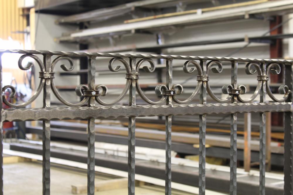 Surowy, metalowy fragment balustrady z kutymi zdobieniami w warsztacie, widoczne ślady obróbki i spawania.