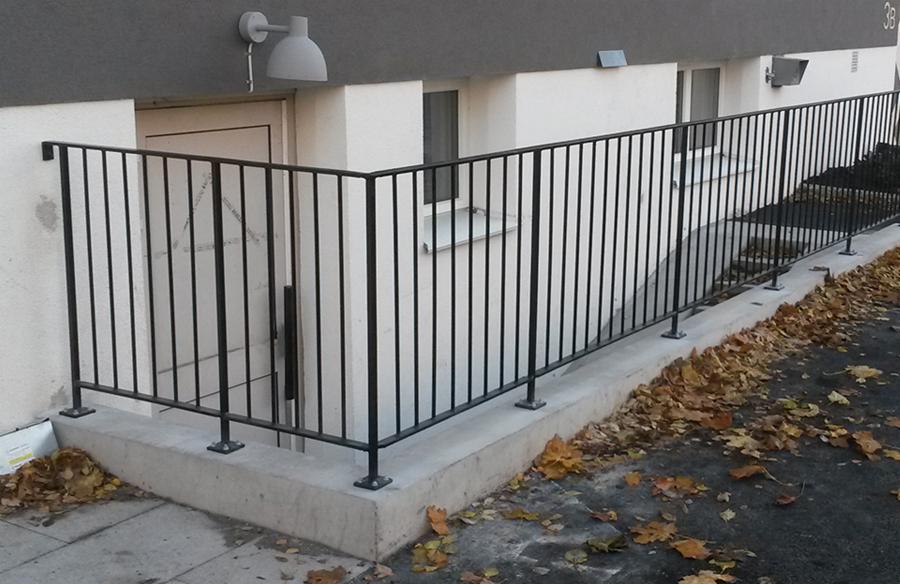 Czarne, metalowe barierki zabezpieczające zejście do piwnicy budynku, widoczne mocowania do betonowej podstawy, jesienne liście na chodniku.