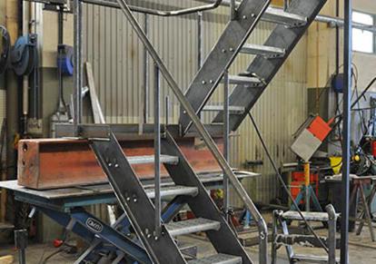 Metalowe schody przemysłowe z poręczami w surowym warsztacie, widoczne ślady spawania i obróbki, na tle blaszane ściany i narzędzia.