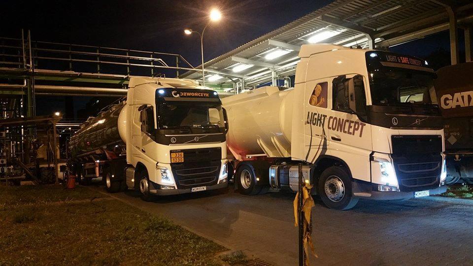 Dwa białe ciągniki siodłowe z cysternami, jeden z napisem 'Light Concept' i grafiką, drugi z logo 'Dzwonczyk', stojące na tle przemysłowej instalacji oświetlonej lampami ulicznymi.