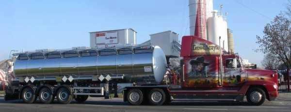 Czerwona ciężarówka z chromowaną cysterną i malowidłami na kabinie, zaparkowana na tle silosów.