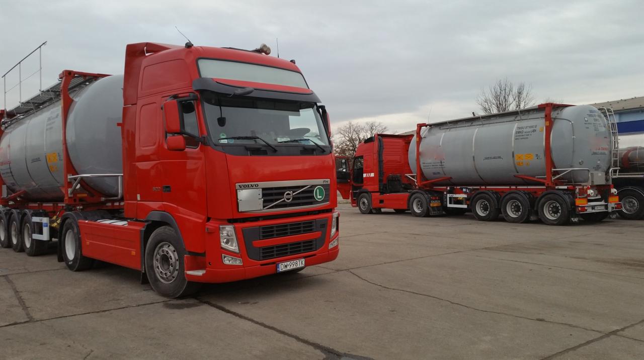 Czerwona ciężarówka Volvo FH z szarą cysterną na parkingu, widoczne inne podobne zestawy, pochmurne niebo w tle.