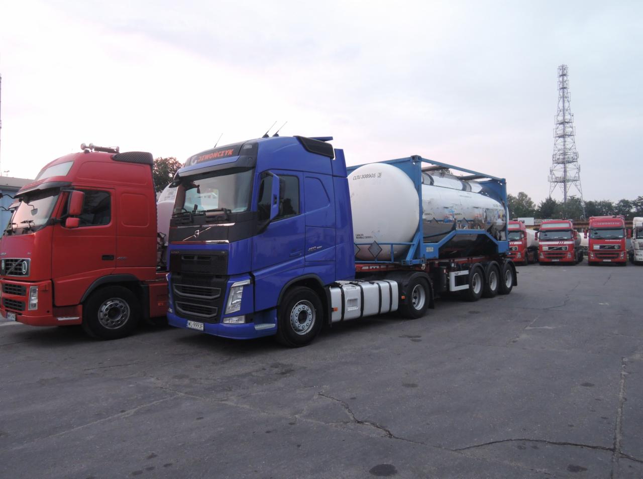 Ciężarówka Volvo w kolorze niebieskim z cysterną na naczepie, zaparkowana obok czerwonej ciężarówki Volvo na betonowym placu, w tle inne ciężarówki i wysoka wieża telekomunikacyjna.