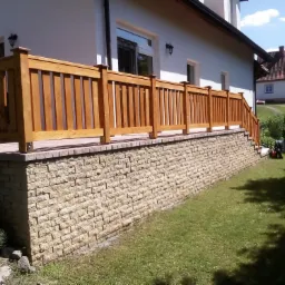 Drewniana balustrada tarasowa z pionowymi szczeblami, zamontowana na murku z kamienia łupanego, prowadząca do schodów, z widokiem na trawnik i dom w tle.
