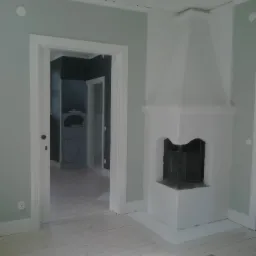 Jasne wnętrze z pomalowanymi na szaro-zielono ścianami, białymi drzwiami i drewnianą podłogą, widoczny biały, narożny kominek z metalowymi drzwiczkami oraz widok na kolejne pomieszczenie z szarą...