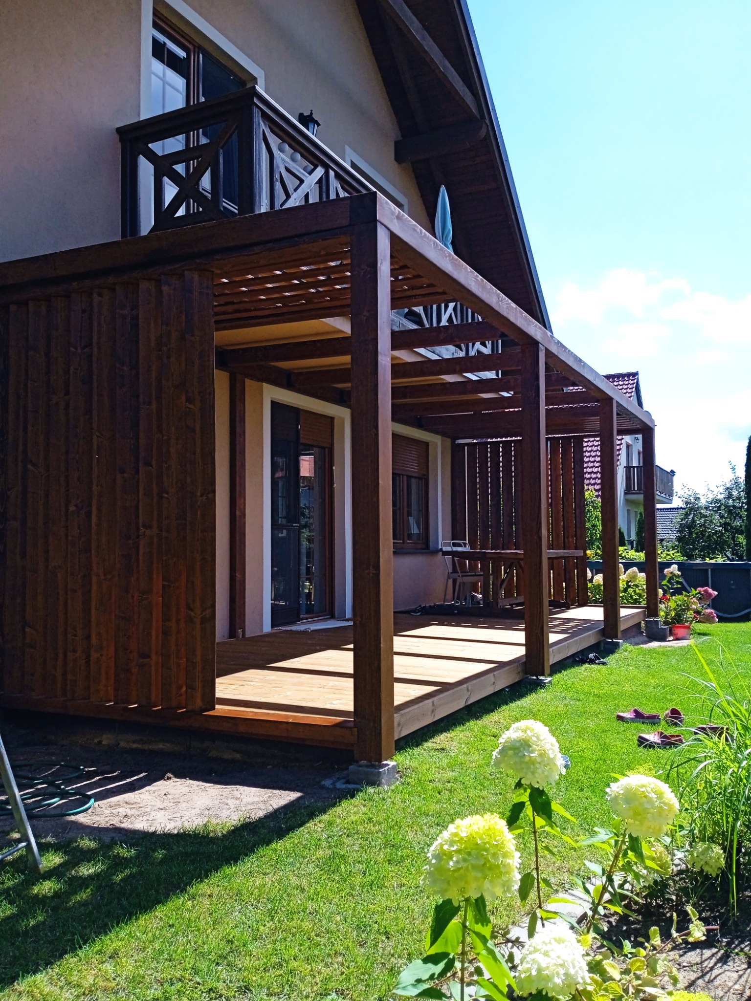 Nowoczesna, ciemnobrązowa konstrukcja z drewna, częściowo zabudowana, z drewnianym podestem, przylegająca do domu, z widocznymi promieniami słońca na trawniku i kwitnącymi hortensjami w pierwszym...
