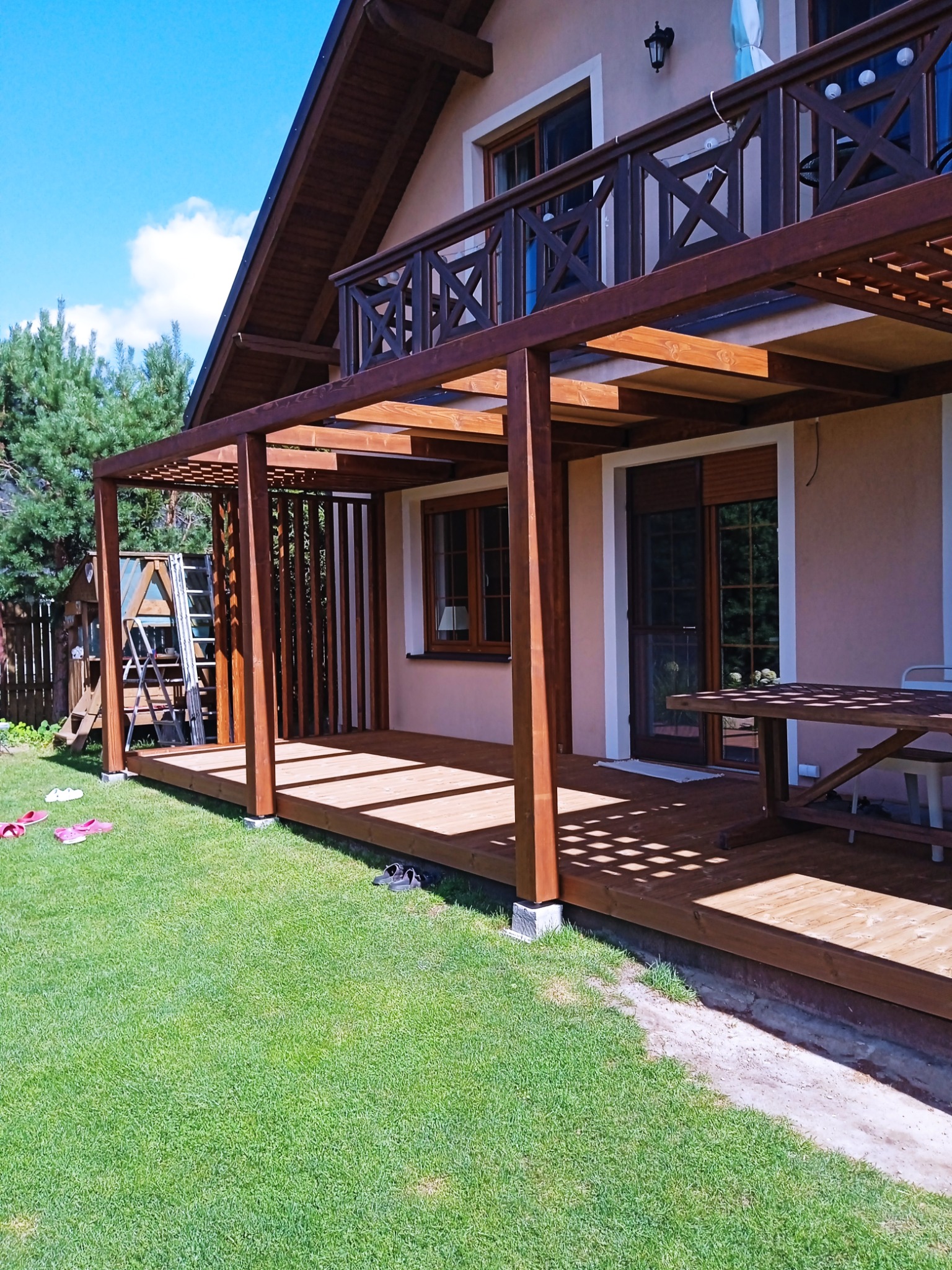 Drewniany taras z pergolą przylegający do domu, z widocznymi elementami konstrukcyjnymi i zacienieniem, na trawniku leżą buty, w tle drabina oparta o budynek.