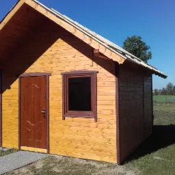 Mała, drewniana konstrukcja z brązowymi drzwiami i oknem na tle zieleni i błękitnego nieba; widoczne elementy konstrukcyjne dachu.