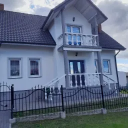 Wykończona elewacja domu jednorodzinnego z balkonem i czarną, metalową bramą wjazdową na tle pochmurnego nieba.