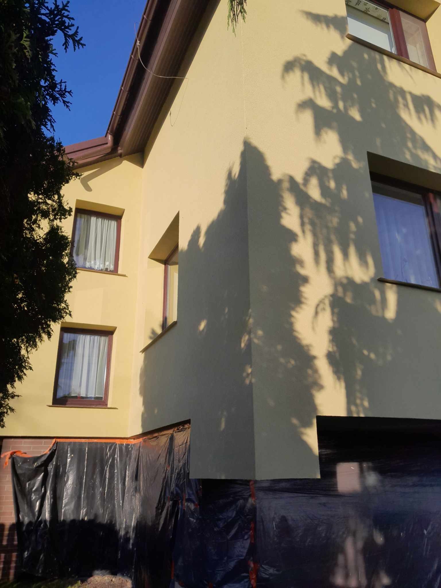 Żółta elewacja budynku z brązowymi oknami, częściowo zasłonięta czarną folią ochronną, cień drzewa na ścianie