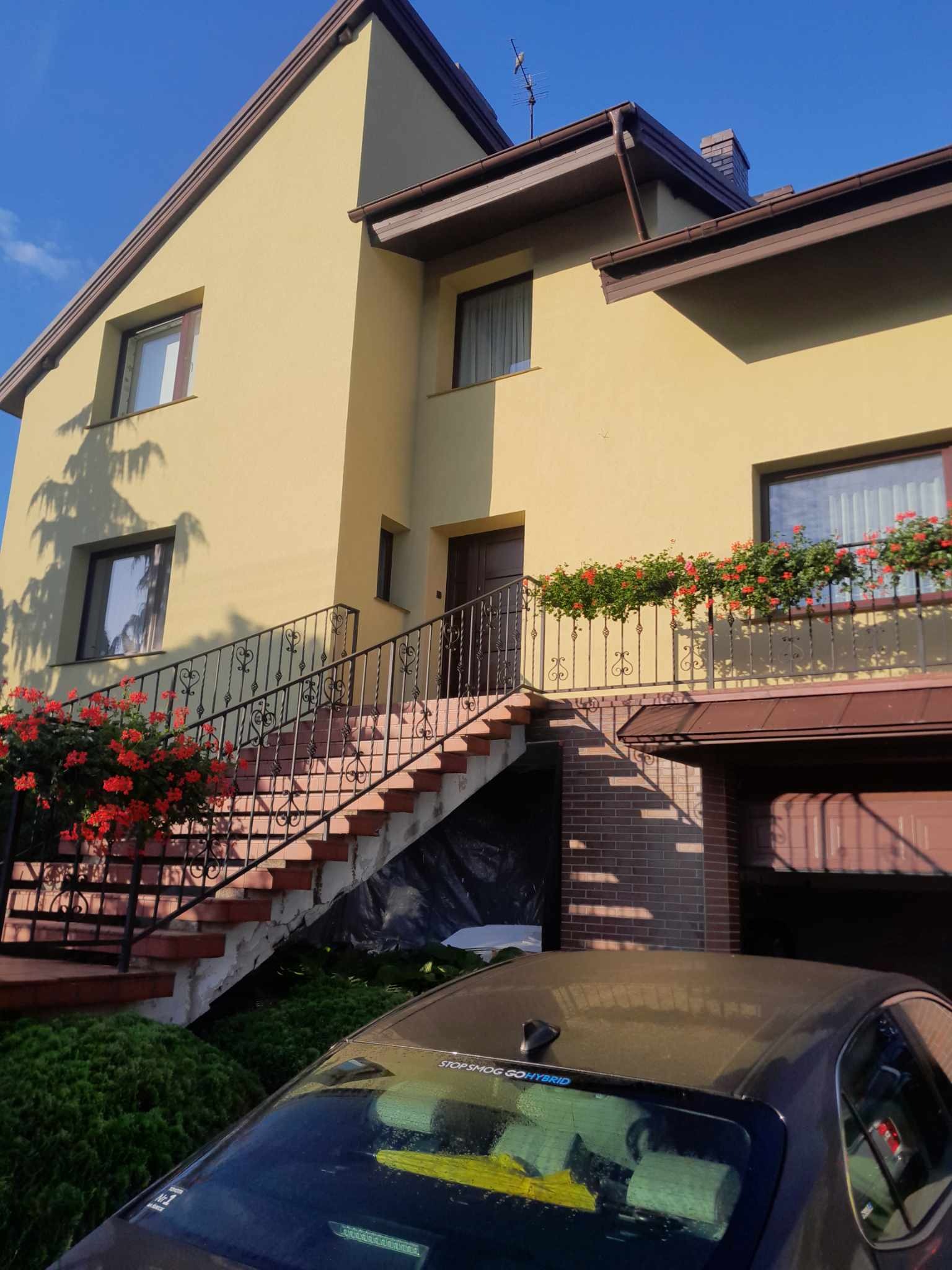 Żółta elewacja dwupiętrowego domu z brązowymi detalami, schody wejściowe z kutą balustradą ozdobioną czerwonymi kwiatami, częściowo zasłonięte auto na pierwszym planie.