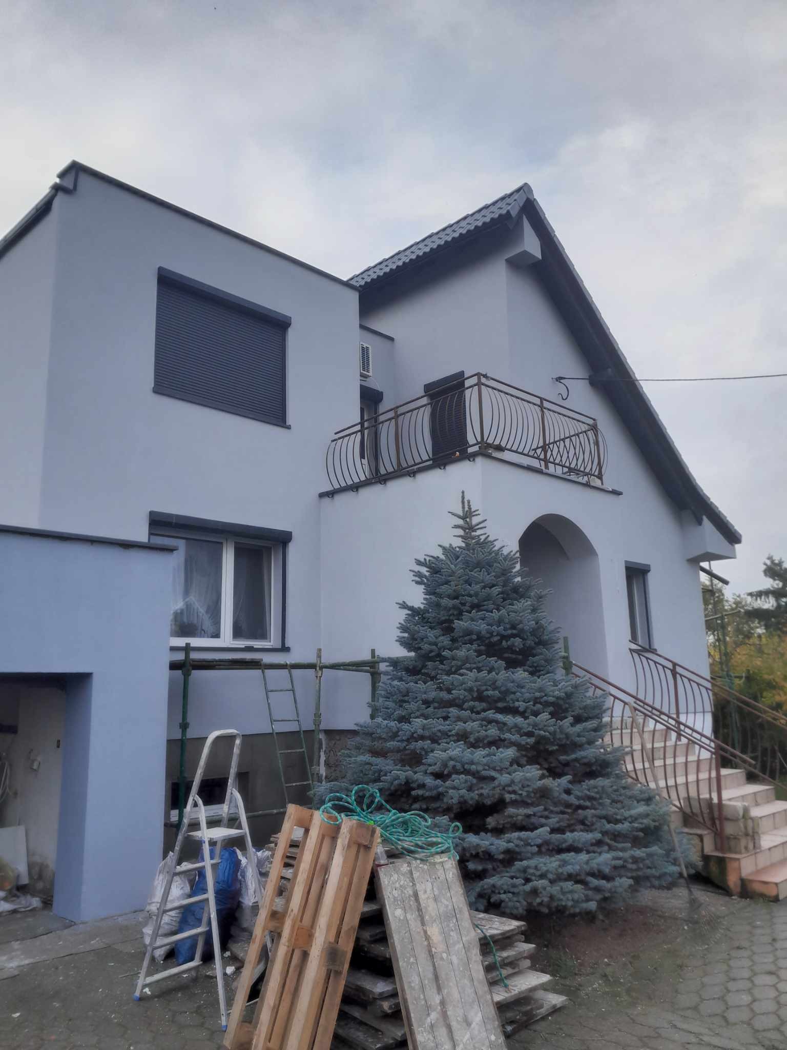 Dwukondygnacyjny dom z oknami z roletami i balkonem z kutą balustradą, świeżo pomalowany na szaro, z rusztowaniem i drewnianymi paletami u podnóża