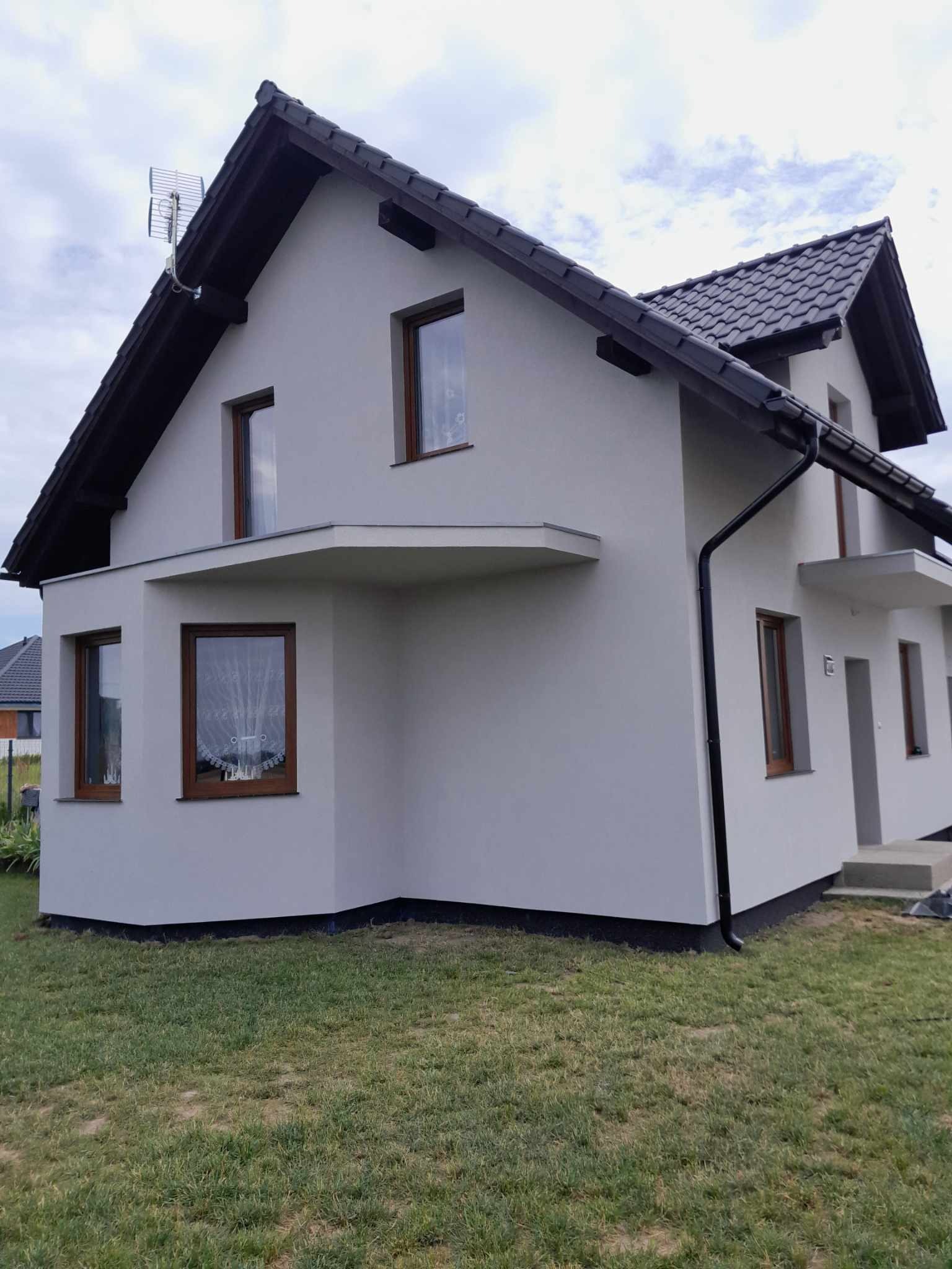 Jasnoszary dom jednorodzinny z ciemnobrązową dachówką, widok z trawnika, antena telewizyjna na dachu.