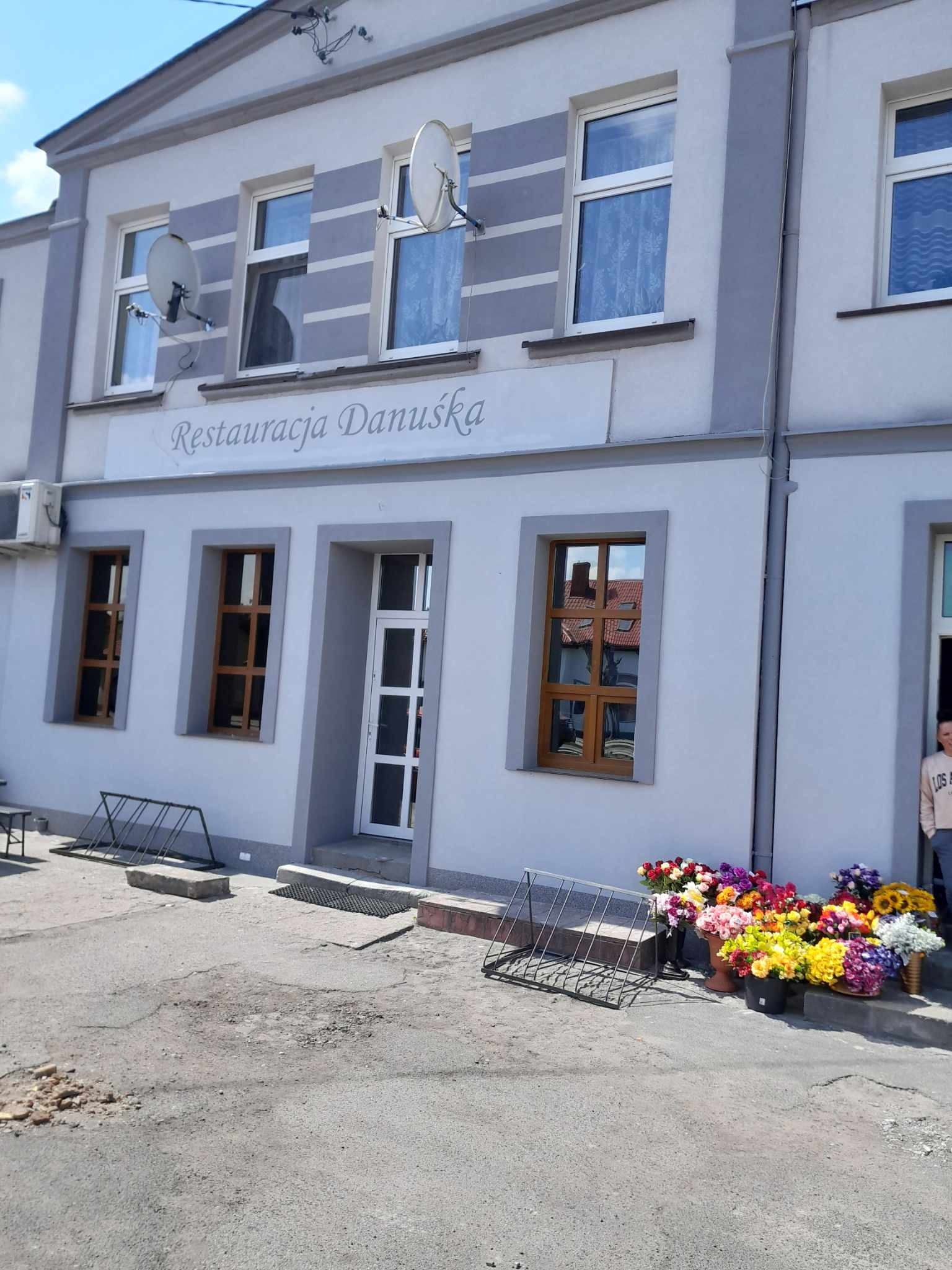 Dwupiętrowy budynek restauracji z szarą elewacją, brązowymi ramami okiennymi i białymi drzwiami wejściowymi, z szyldem 'Restauracja Danuśka' nad wejściem, satelitami na górze i kwiatami w donicach...
