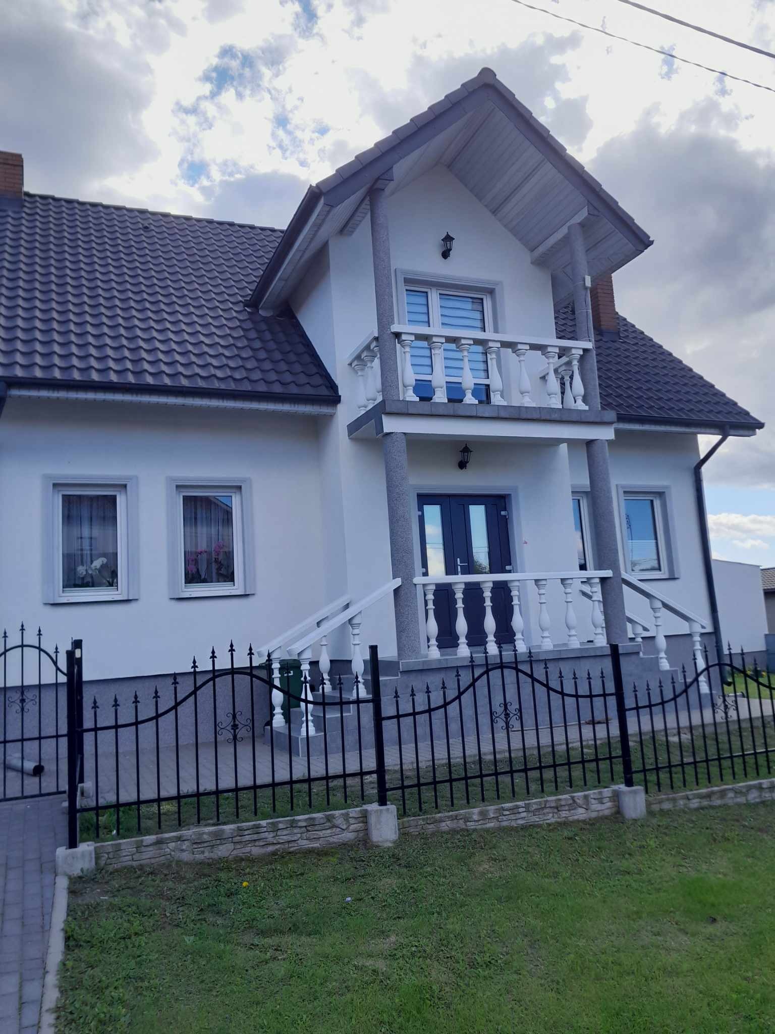 Wykończona elewacja domu jednorodzinnego z balkonem i czarną, metalową bramą wjazdową na tle pochmurnego nieba.