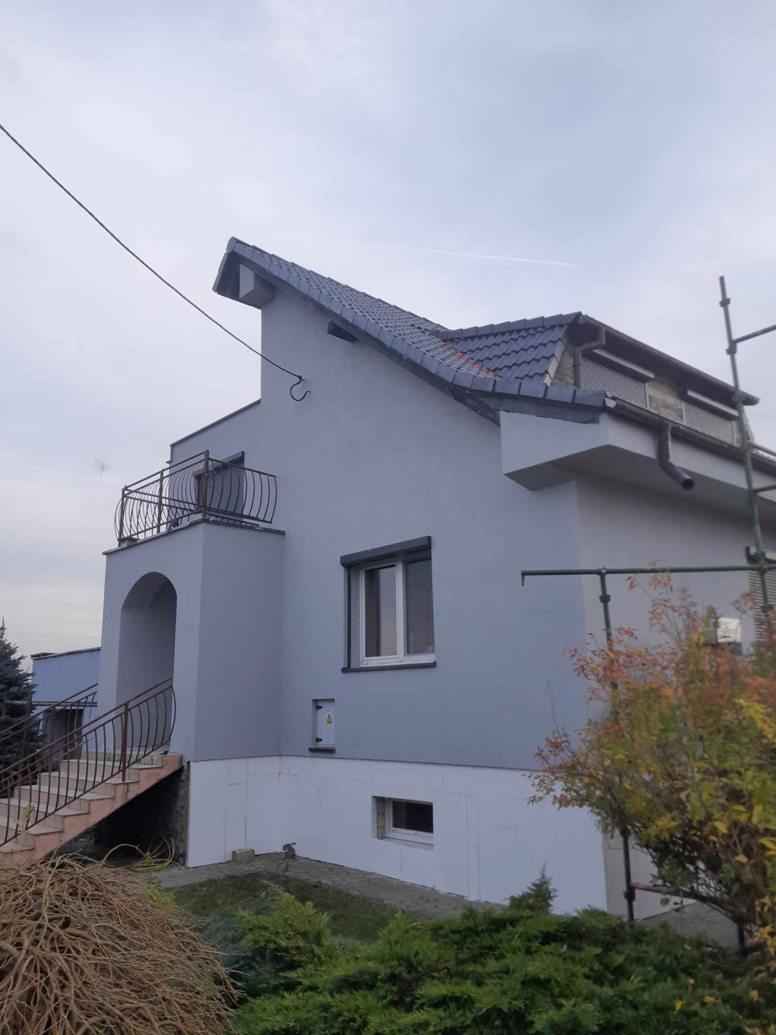Dom jednorodzinny po remoncie elewacji, widoczne ocieplenie styropianem i świeżo pomalowana szara fasada, balkon z kutą balustradą, fragment rusztowania po prawej stronie.