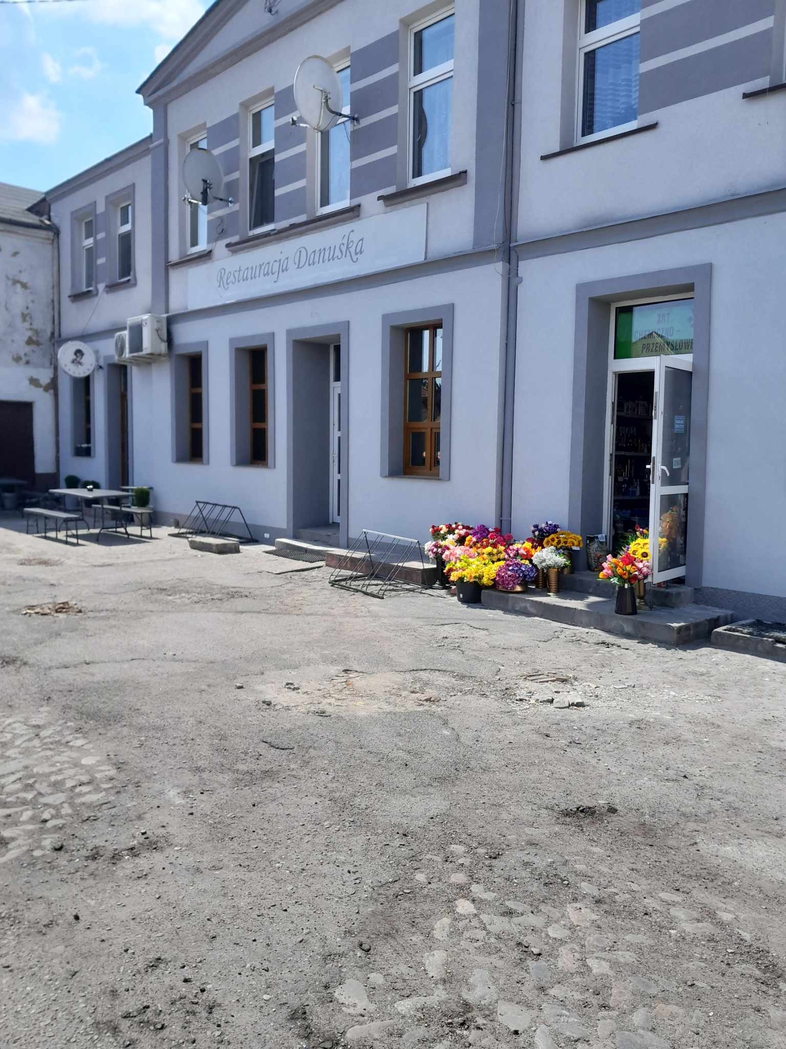 Jasna fasada budynku z szarymi akcentami, widoczne okna z brązowymi ramami, napisem 'Restauracja Danuśka' oraz bukietami kolorowych kwiatów w donicach przed wejściem.