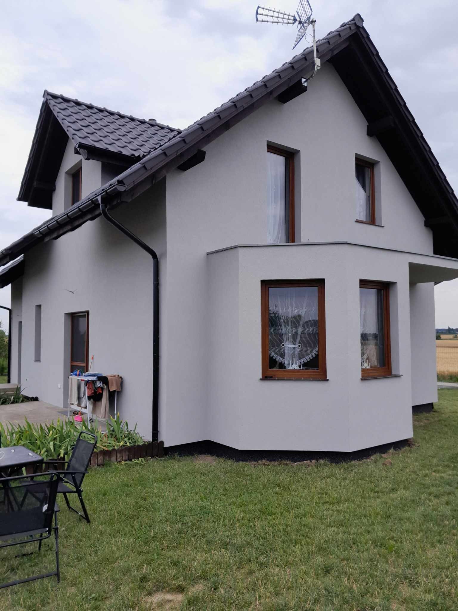 Dom jednorodzinny z szarą elewacją, brązowymi oknami i ciemnym dachem, antena telewizyjna na dachu, widok z trawnika.