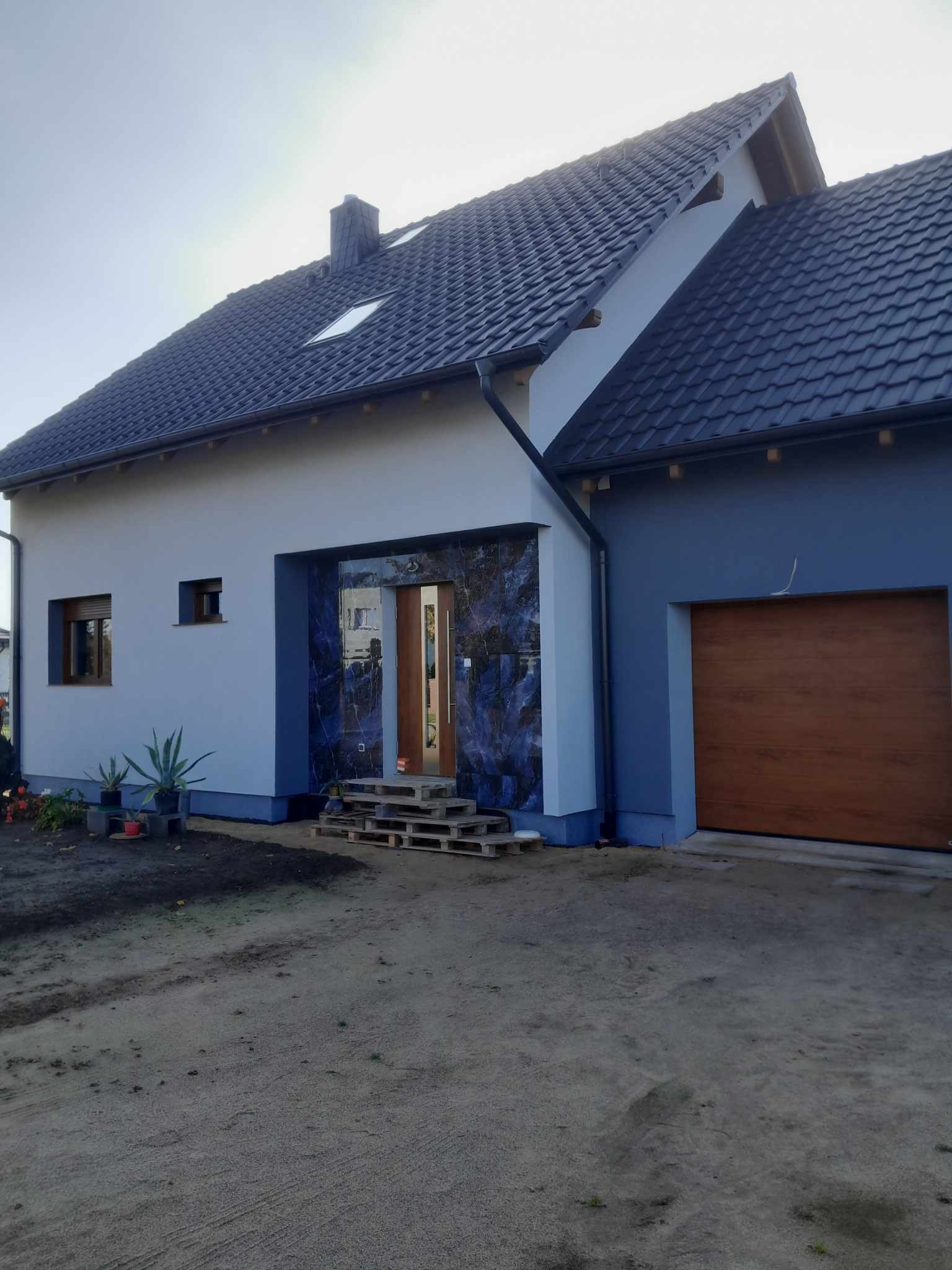 Nowoczesny dom jednorodzinny z elewacją w odcieniach szarości i niebieskiego, wejście wykończone płytami imitującymi marmur, drewniana brama garażowa, palety przed wejściem.