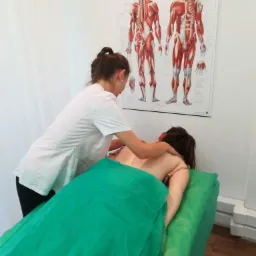 Kobieta w białym uniformie wykonuje masaż pleców pacjentki leżącej na zielonym stole do masażu, w tle plakat z układem mięśni człowieka.