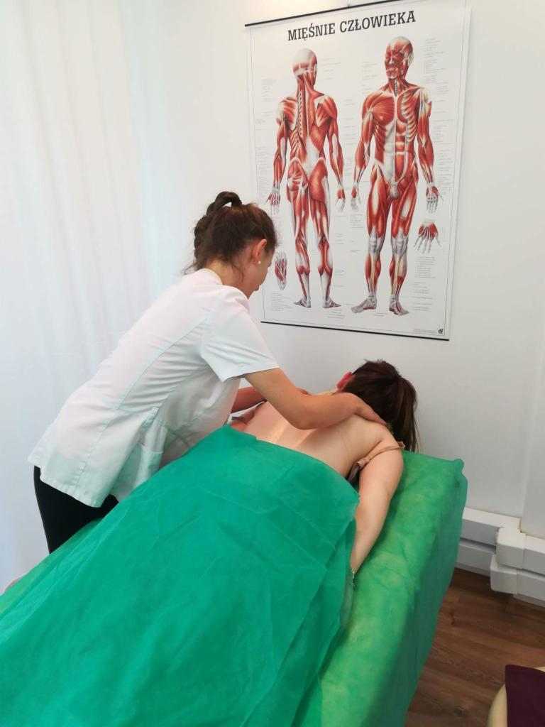 Kobieta w białym uniformie wykonuje masaż pleców pacjentki leżącej na zielonym stole do masażu, w tle plakat z układem mięśni człowieka.