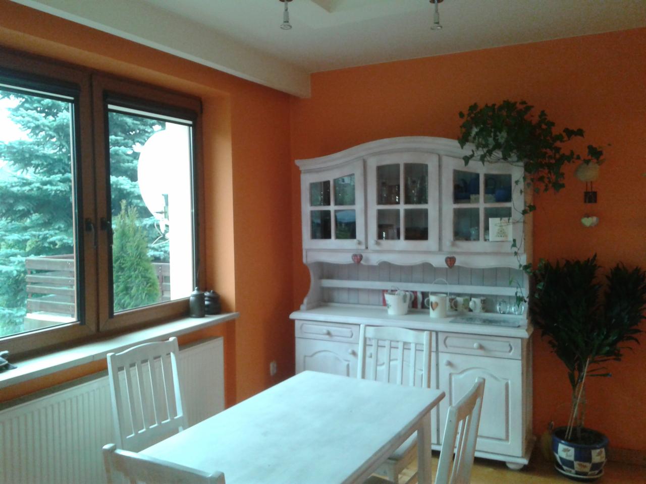 Jasna jadalnia z pomarańczowymi ścianami, białym kredensem i stołem. Za oknem widok na iglaste drzewa.