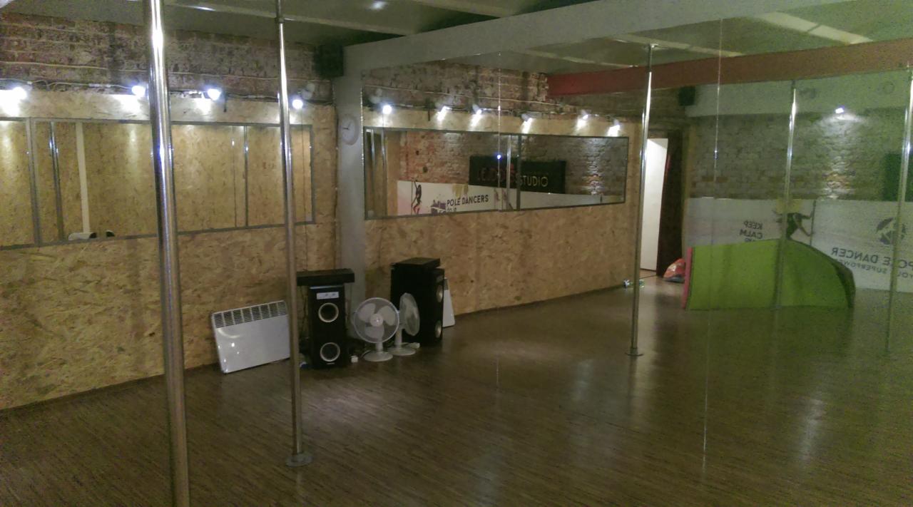Puste studio tańca z lustrami, parkietem, grzejnikiem, wentylatorami i plakatem 'Pole Dancers' na ścianie.