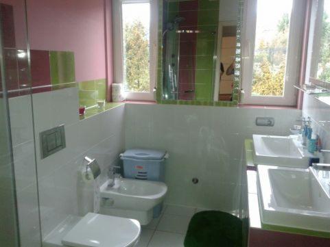 Łazienka z dwoma umywalkami, bidetem i toaletą w jasnych kolorach z akcentami różu i zieleni. Na ścianie nad umywalkami widoczne okna z widokiem na drzewa.