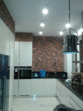 Nowoczesna kuchnia z białymi szafkami i ścianą z czerwonej cegły, oświetlona punktowymi lampami sufitowymi i wiszącą lampą nad blatem. Widoczny niebieski pojemnik na blacie.